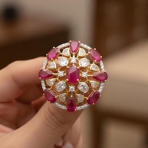 May include: A gold-toned ring featuring a central oval ruby surrounded by pear-shaped rubies and round, clear gemstones. The ring has a circular design with a halo of small, clear stones around the outer edge.
