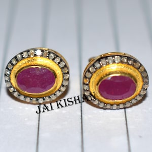 May include: A pair of oval-shaped earrings with a red ruby gemstone set in a gold-toned metal frame. The frame is surrounded by a halo of small, clear diamonds.