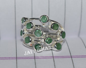 May include: A silver ring with multiple bands set with green gemstones. The ring is on a white and purple lined surface.