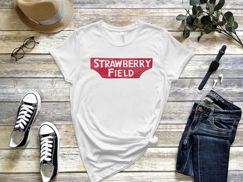 May include: White t-shirt with a red vintage-style sign that says "STRAWBERRY FIELD" printed on the front.