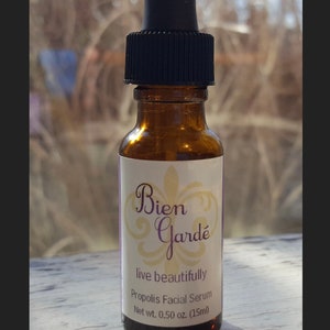 May include: A small amber glass bottle with a black dropper top. The bottle is labeled "Bien Gard&eacute;" with a decorative fleur-de-lis design. The label also reads "live beautifully" and "Propolis Facial Serum" with a net weight of 0.50 oz (15 ml).