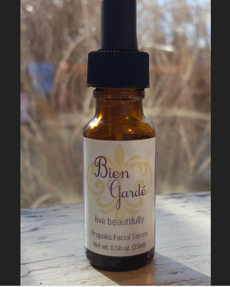 May include: A small amber glass bottle with a black dropper top. The bottle is labeled "Bien Gard&eacute;" with a decorative fleur-de-lis design. The label also reads "live beautifully" and "Propolis Facial Serum" with a net weight of 0.50 oz (15 ml).