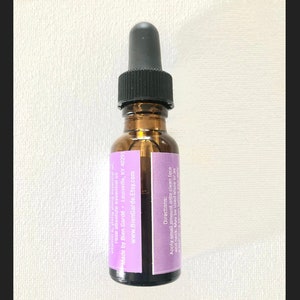 May include: A small glass bottle with a dropper top, filled with a clear liquid. The bottle has a purple label with white text that reads "vitamin e, ylang ylang essential oil, rose absolute essential oil, Made by Bien Gard&eacute; - Louisville, KY 40291, www.BienGarde.Etsy.com, Directions: Apply small amount onto clean face and neck. May be used alone or as a hydrating serum before your daily".