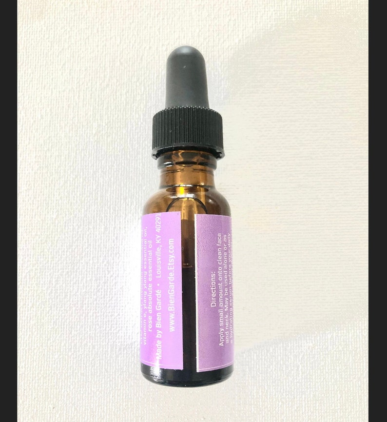 May include: A small glass bottle with a dropper top, filled with a clear liquid. The bottle has a purple label with white text that reads "vitamin e, ylang ylang essential oil, rose absolute essential oil, Made by Bien Gard&eacute; - Louisville, KY 40291, www.BienGarde.Etsy.com, Directions: Apply small amount onto clean face and neck. May be used alone or as a hydrating serum before your daily".