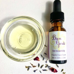 May include: A small amber glass bottle with a dropper top, containing a clear liquid. The bottle is labeled "Bien Gard&eacute; live beautifully Propolis Facial Serum Net wt. 0.50 oz. (15ml)". The bottle is surrounded by dried lavender flowers and rose petals on a white surface.