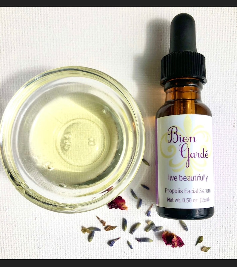 May include: A small amber glass bottle with a dropper top, containing a clear liquid. The bottle is labeled "Bien Gard&eacute; live beautifully Propolis Facial Serum Net wt. 0.50 oz. (15ml)". The bottle is surrounded by dried lavender flowers and rose petals on a white surface.