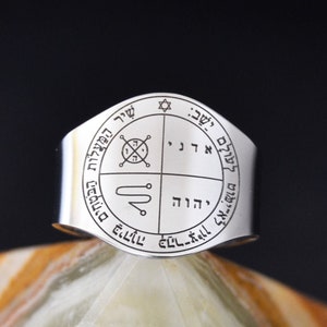 May include: A silver ring with a circular design featuring Hebrew text and symbols. The ring is on a light brown and white marble surface.