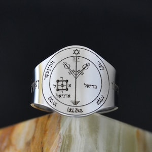May include: A silver ring with a sigil engraved on the top. The sigil is surrounded by Hebrew text. The ring is sitting on a brown and white stone.