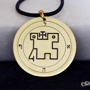 May include: A gold-colored pendant necklace with a black cord. The pendant features a circular design with a geometric symbol and Hebrew lettering.