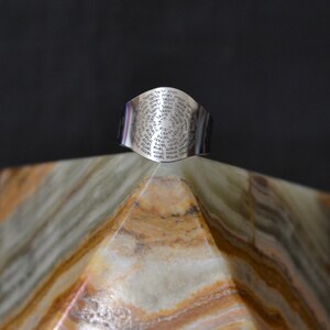 May include: A silver ring with a circular design featuring a repeating pattern of text. The ring is sitting on a pyramid-shaped piece of marble.