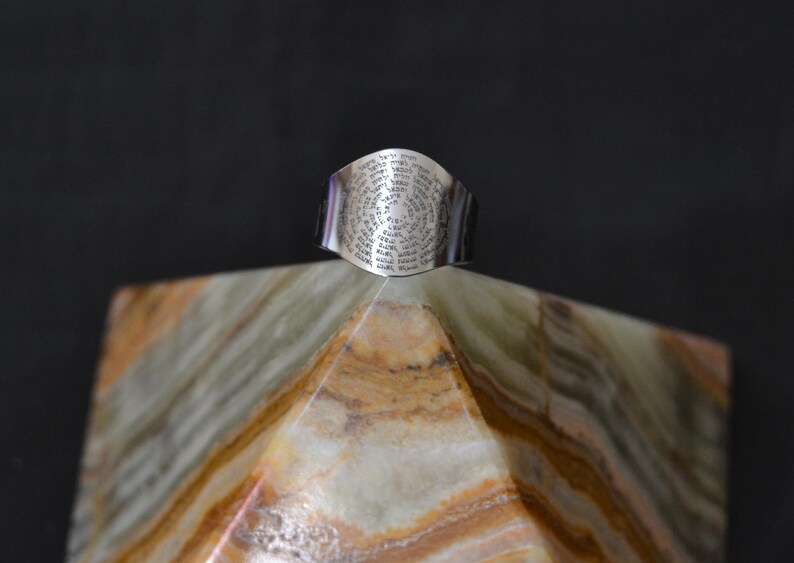 May include: A silver ring with a circular design featuring a repeating pattern of text. The ring is sitting on a pyramid-shaped piece of marble.