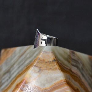 May include: A silver ring with a geometric design and the letter "J" cut out of the band. The ring is sitting on a white and brown marble pyramid.