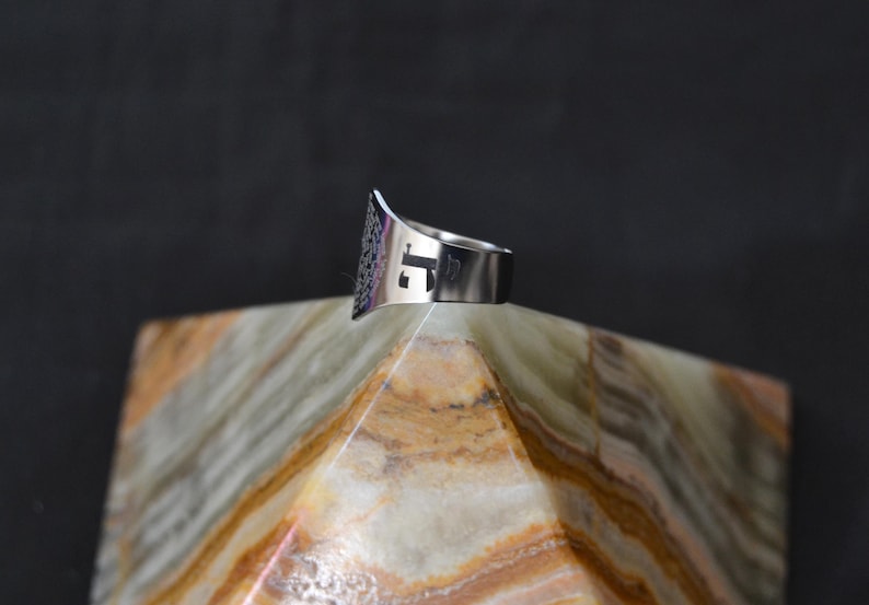 May include: A silver ring with a geometric design and the letter "J" cut out of the band. The ring is sitting on a white and brown marble pyramid.