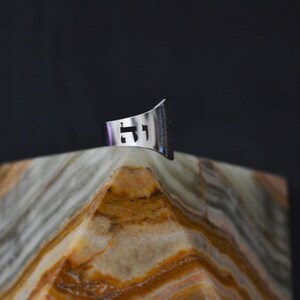 May include: A silver ring with a cut-out design of the Hebrew letters "Chai" on a textured, light brown surface.