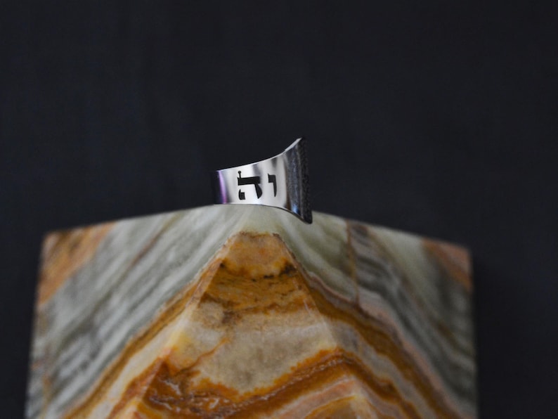 May include: A silver ring with a cut-out design of the Hebrew letters "Chai" on a textured, light brown surface.