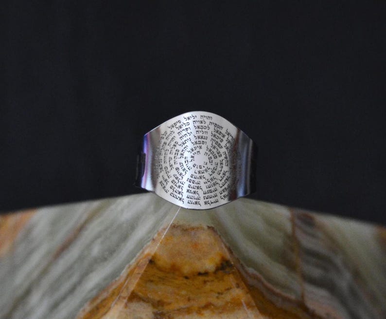 May include: A silver ring with a circular design featuring Hebrew text. The ring is on a light green and brown surface.