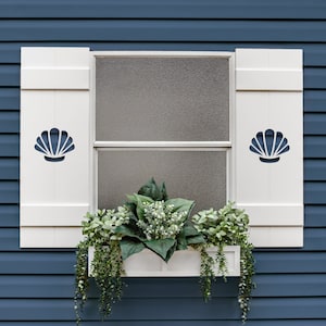 May include: A white window with matching shutters featuring a seashell design. The window box is filled with greenery and hanging plants. The window is set against a blue exterior wall.
