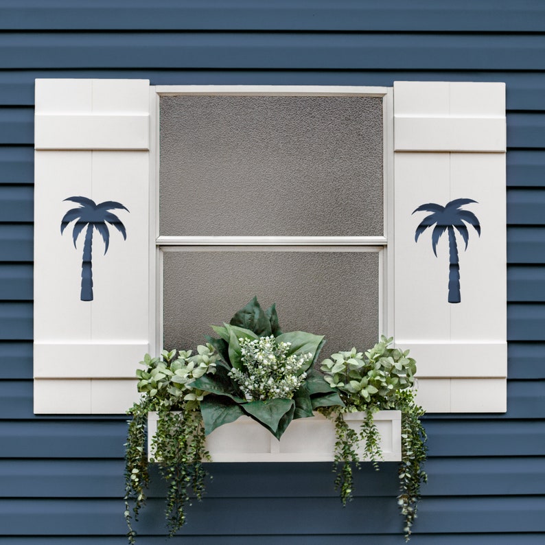 May include: White window shutters with a dark blue palm tree cutout design. A white window box below the window holds green and white plants. The window is set against a blue exterior wall.