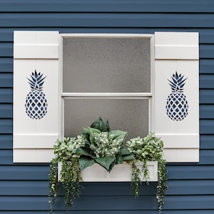 May include: White window shutters with a pineapple design, a white window box planter with green plants, and a blue siding background.