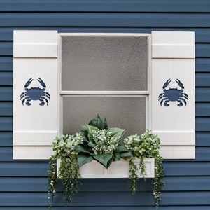 May include: White window shutters with a blue crab cutout design. The shutters flank a window with a white frame and a white window box filled with greenery and hanging plants. The background is a blue wall.