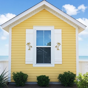 May include: A small, yellow beach house with white trim and a black roof. The house has a window with white shutters and palm tree decals. Green bushes are planted in front of the house, and the ocean is visible in the background.