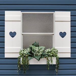 May include: White window shutters with heart cutouts, a window box planter with green plants, and a blue siding background.