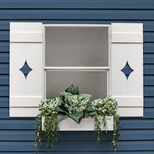 May include: A white window with matching shutters and a diamond-shaped cutout. The window box is filled with greenery and white flowers, with trailing vines. The background is a blue siding.