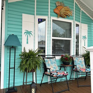 May include: A turquoise exterior with white shutters featuring palm tree cutouts. A metal fish decorates the wall above a window. The porch is furnished with black chairs, floral cushions, and potted plants, creating a tropical theme.