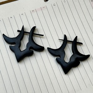 May include: A pair of black wooden earrings with a unique abstract design. The earrings are shaped like a stylised crescent moon with a small cutout in the centre.