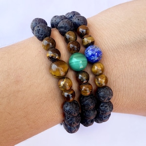 May include: Three bracelets stacked on a wrist. The bracelets are made of black lava rock beads and tiger's eye beads. The top bracelet has a blue lapis lazuli bead and a green malachite bead.