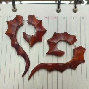 May include: Two wooden earrings with a unique, intricate design. The earrings are a deep reddish-brown colour and have a curved shape with sharp points.