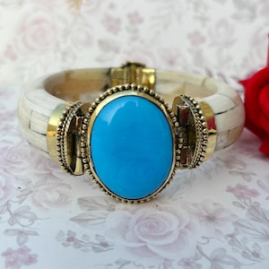 May include: Ivory-coloured bangle bracelet featuring a large, oval turquoise stone set in a gold-coloured metal frame. The bracelet has gold-coloured metal accents and a hinged clasp. The bracelet is a statement piece.