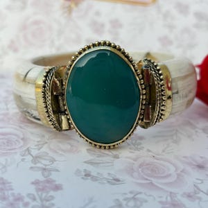May include: A wide bracelet featuring a large, oval, emerald-green stone set in a gold-coloured metal frame. The bracelet has a cream-coloured band with gold accents and decorative metalwork. A statement piece of jewellery.
