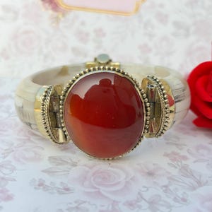 May include: A wide, cream-coloured bangle bracelet featuring a large, oval, red-orange stone at its centre. The stone is set in a gold-coloured metal frame. The bracelet has a hinged clasp and decorative metal accents.