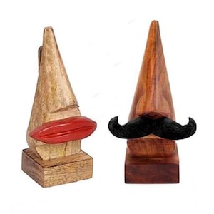 Puede incluir: Dos esculturas de madera de caras. Una tiene un labio rojo y la otra un bigote negro. Ambas están sobre una base cuadrada y tienen una cabeza en forma de cono. Las esculturas están hechas de madera con diferentes tonos de marrón.