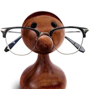May include: A wooden eyeglasses holder in the shape of a face, with black framed glasses resting on the nose. The holder is made of dark brown wood and has a smooth finish. The glasses have a silver bridge and clear lenses.