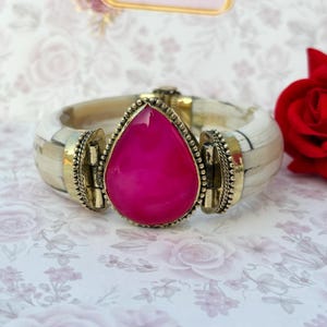 May include: A chunky bracelet with a vibrant pink, teardrop-shaped gemstone set in a gold-coloured metal frame. The bracelet band is cream with a mosaic pattern. A red rose is in the background.