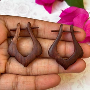 May include: A pair of dark brown wooden earrings with a unique teardrop shape. The earrings are held together by a small metal pin.