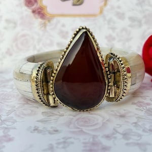 May include: A wide, cream-coloured bracelet featuring a large, teardrop-shaped, dark red stone at the centre. The stone is framed by a gold-coloured metal setting with a beaded edge. The bracelet has gold-coloured metal accents.