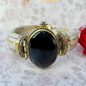 May include: A wide, ivory-coloured bangle bracelet featuring a large, oval black stone centrepiece. The stone is set in a gold-toned metal frame, and the bracelet has gold accents. The bracelet has a hinged clasp.