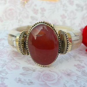 May include: A wide bangle bracelet with a large, oval, reddish-brown stone set in a gold-coloured metal frame. The bracelet band is cream-coloured with inlaid bone-like segments. A striking piece of jewellery.