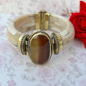 May include: A cream-coloured bangle bracelet with a large, oval, amber-coloured stone set in a gold-toned metal frame. The bracelet has a hinged clasp and is decorated with gold-toned metal accents.