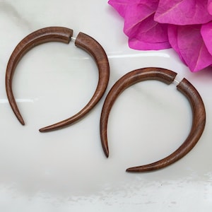 May include: Two dark brown, crescent-shaped wooden earrings. Each earring features a curved design with a silver post. The earrings are presented on a white surface, with a pink flower in the background.