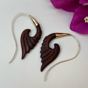 May include: A pair of handcrafted wooden earrings with a wing-like design. The dark brown wood is carved with detailed lines, and the top of each earring has a gold-coloured metal accent. The earrings have silver-coloured ear wires.