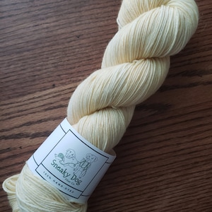 Op de afbeelding: Een bol handgeverfde garen in een lichtgele kleur. Het garen is gewikkeld rond een label met de tekst "Sneaky Dog Fiber Co. 100% Hand-Dyed."