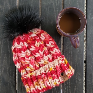 Puede incluir: Un gorro tejido a mano en tonos rojos, blancos y amarillos, rematado con un gran pompón de piel sintética negra. Una etiqueta de cuero marrón está adjunta. Una taza morada llena de café está cerca.