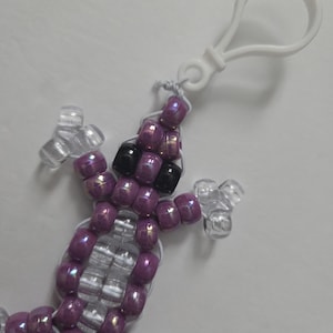 May include: A beaded lizard keychain in shades of purple, clear, and black. The lizard is made of small, round beads strung together with white string. It has a white plastic clip attached for easy attachment to keys or bags.