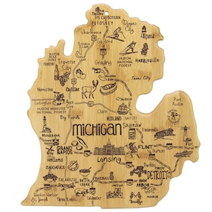 May include: A bamboo cutting board shaped like the state of Michigan. The board features engraved illustrations of cities, landmarks, and icons, including Traverse City, Detroit, and the state capital, Lansing. A hole is drilled at the top for hanging.