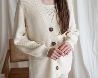May include: A white knitted cardigan with a v-neck, long sleeves, and button closure. The cardigan has a pocket on the left side and is worn over a white lace top.