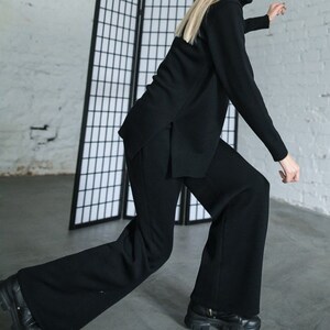 May include: A woman wearing a black turtleneck sweater and black wide-leg pants. She is standing in front of a black room divider.